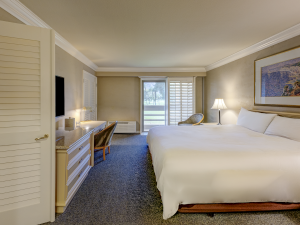 A hotel room with a large bed, desk area, desk chair, nightstands, lamps, a painting, and a balcony view at the far end. 140 characters with a period at the end.