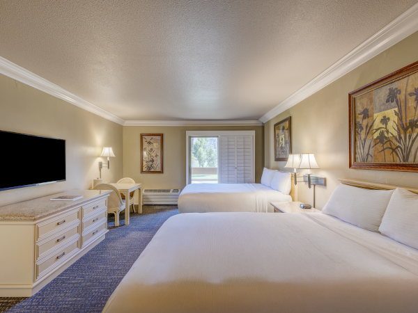A bright hotel room with three white beds, a wall TV, desk, lamps, framed art, and a window at the far end.