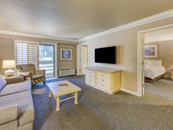 A spacious hotel suite living area with a sofa, armchairs, coffee table, TV on a dresser, and a separate bedroom visible through an open doorway.