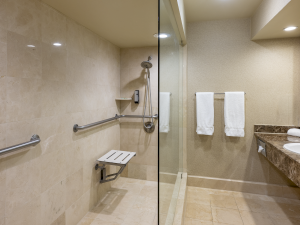 A wheelchair-accessible hotel bathroom with beige tile walls, a walk-in glass shower, grab bars, a fold-down seat, a long vanity with two sinks, and white towels on racks.