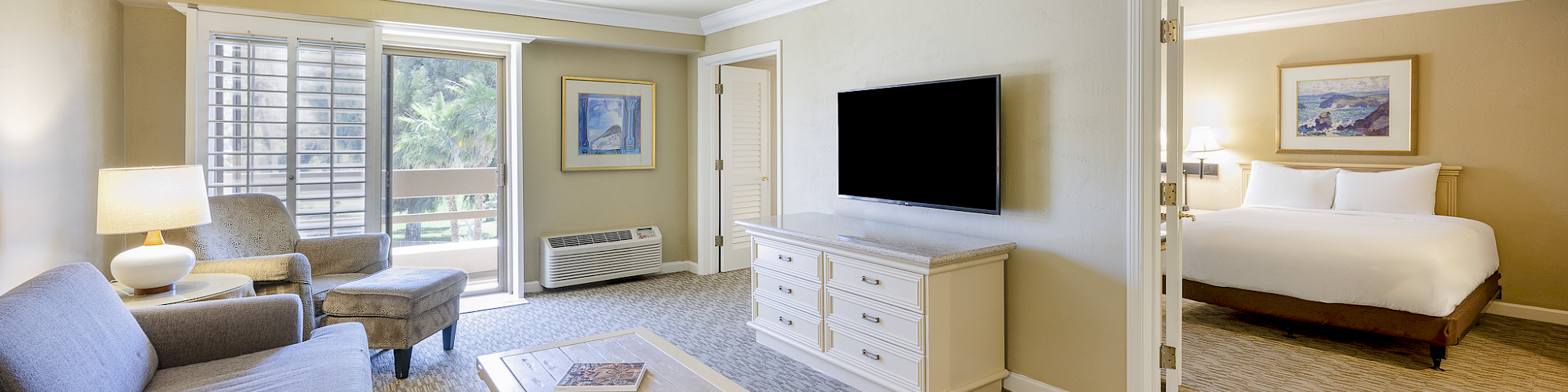 Bright hotel suite living room with couches, coffee table, big wall TV, dresser, lamps, and a glimpse of a bedroom.