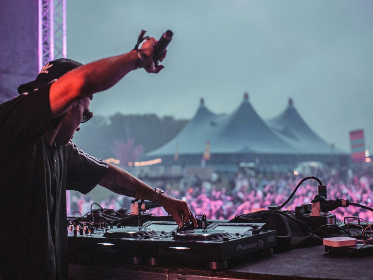 A DJ behind a sound console with one arm raised, performing at a crowded outdoor music festival under colorful lights.