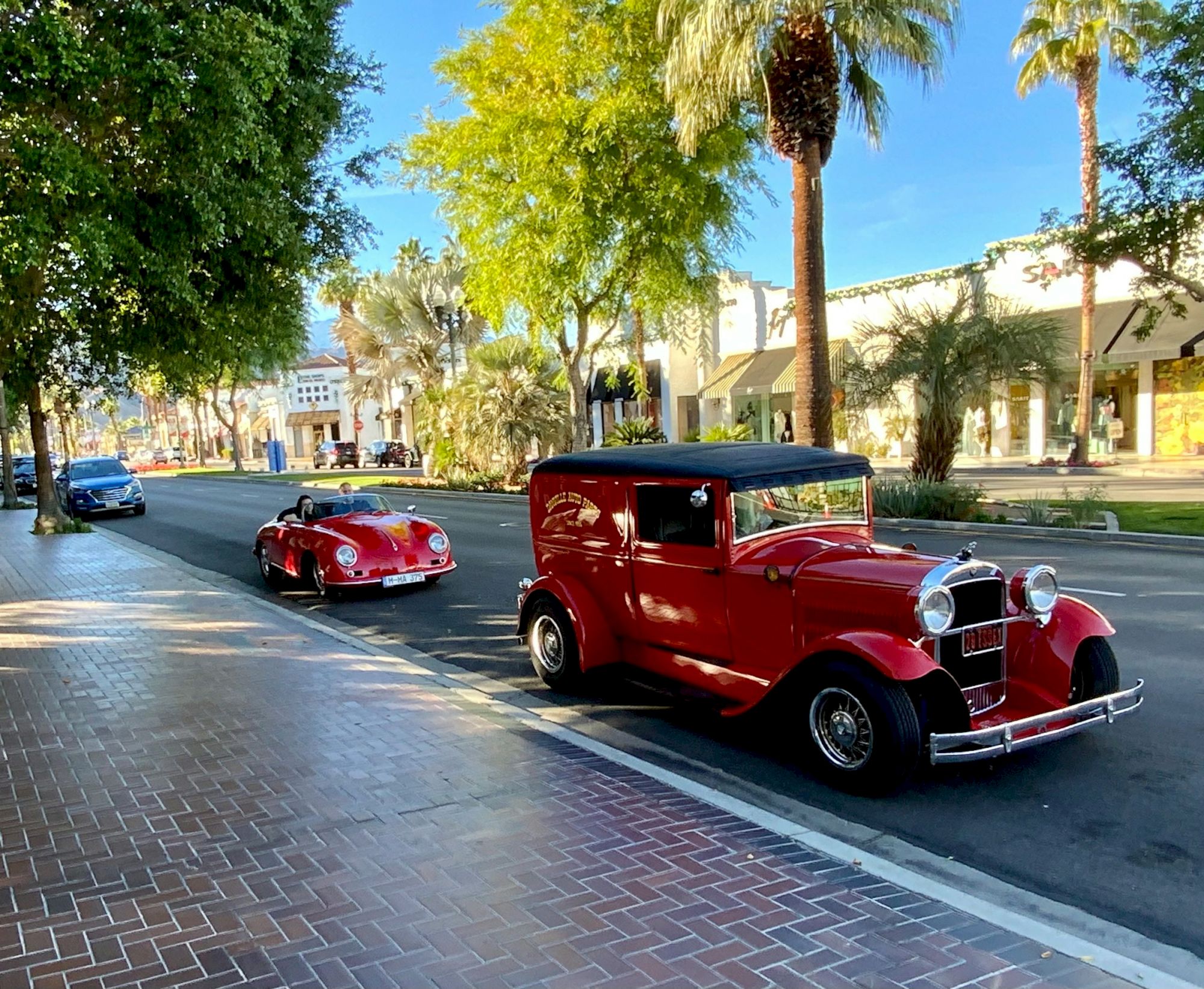 A sunny boulevard lined with palm trees, featuring a red classic car and a vintage bus driving past a resort-like building.
