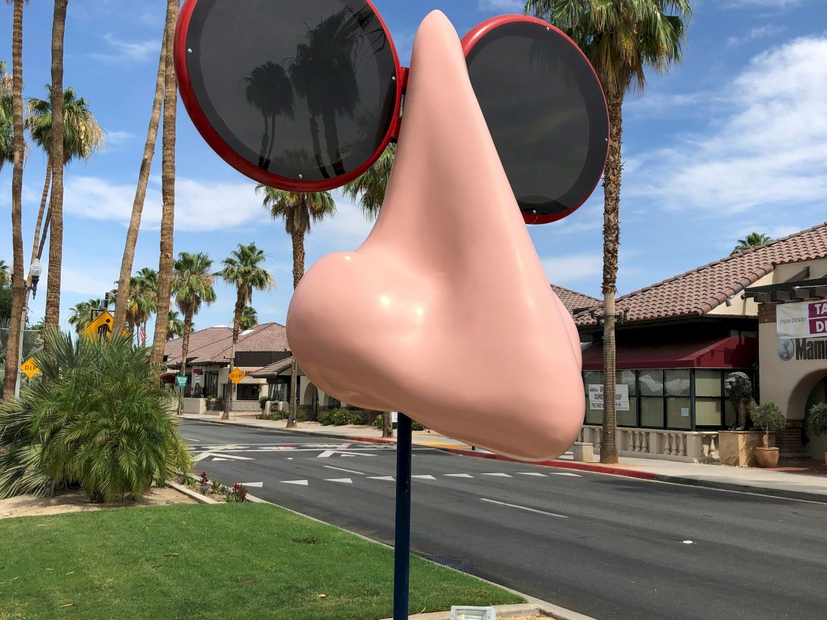 Palm-lined street under a blue sky with a giant cartoon nose and oversized red sunglasses by storefronts, soaking up sunshine.