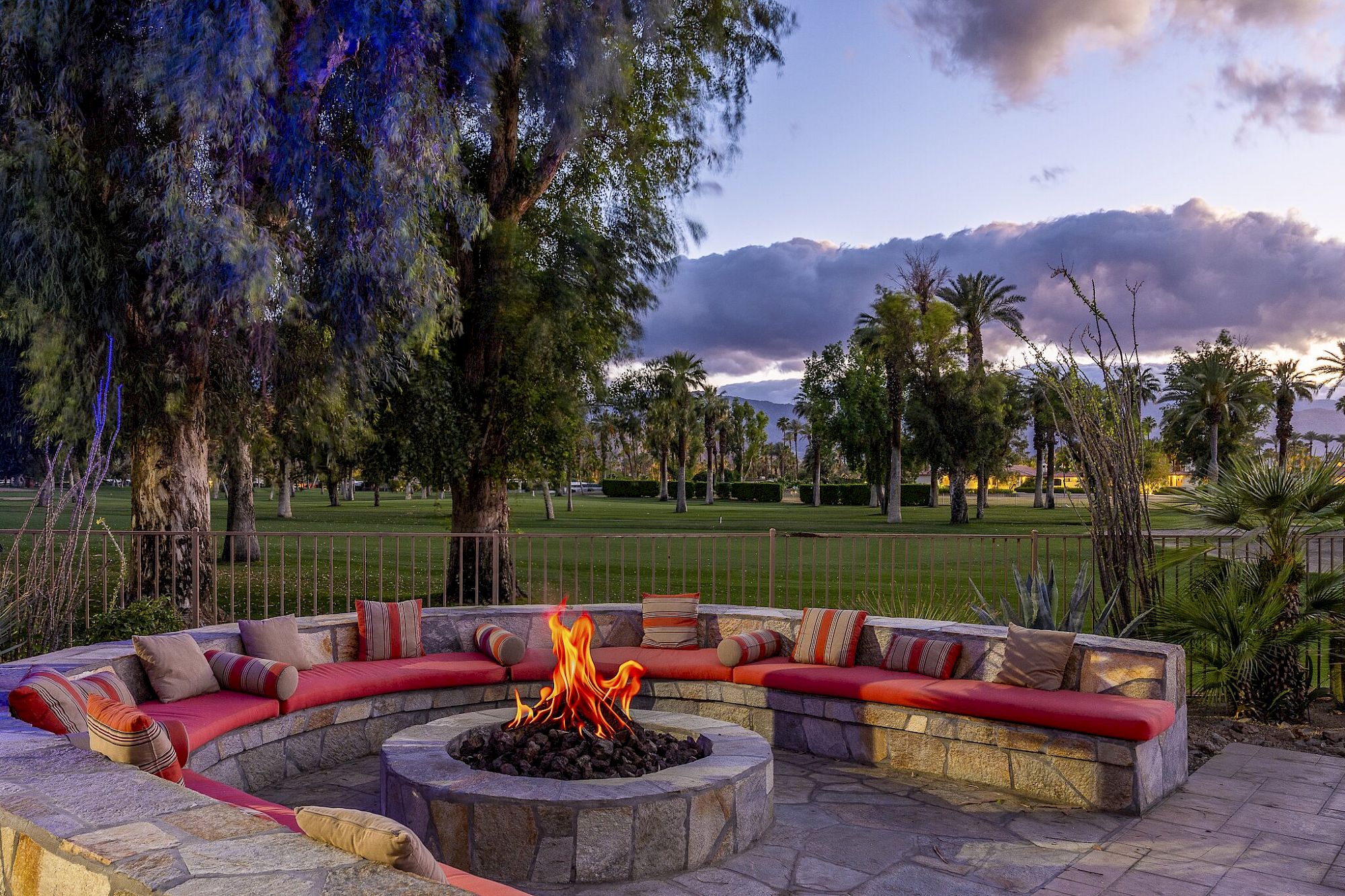 Outdoor patio with a glowing fire pit, red cushioned seating, palm trees, and dramatic mountains under a cloudy dusk sky.