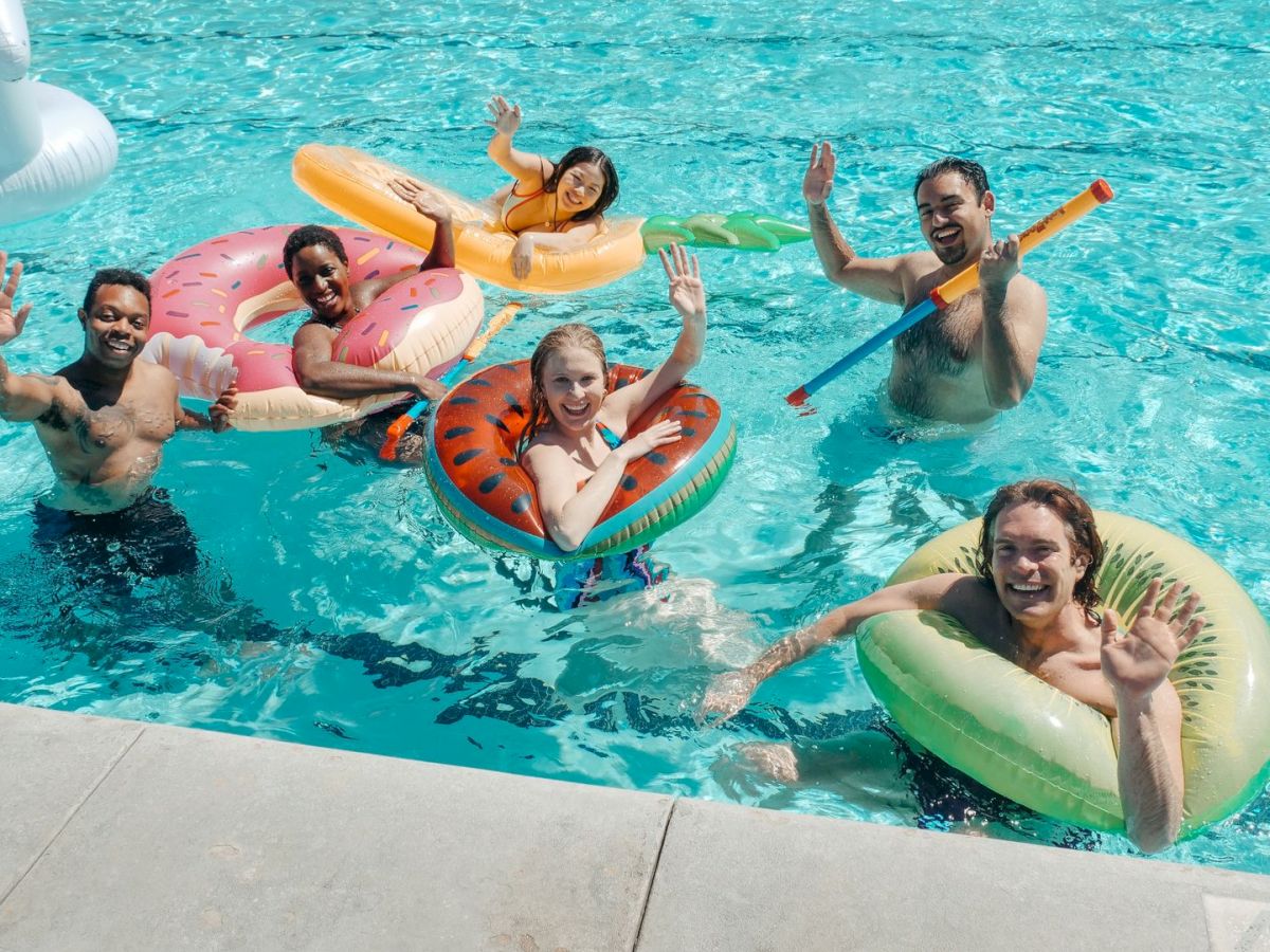 A group of friends and kids splash in a pool, smiling and waving on colorful inflatable rafts and rings.