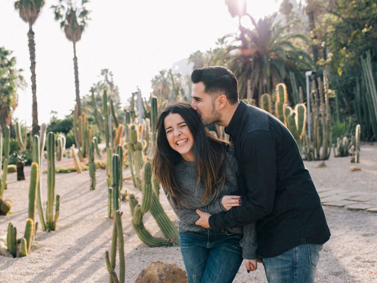 A couple cuddles and smiles in a desert walkway lined with tall cacti, soaking in warm sunset light.