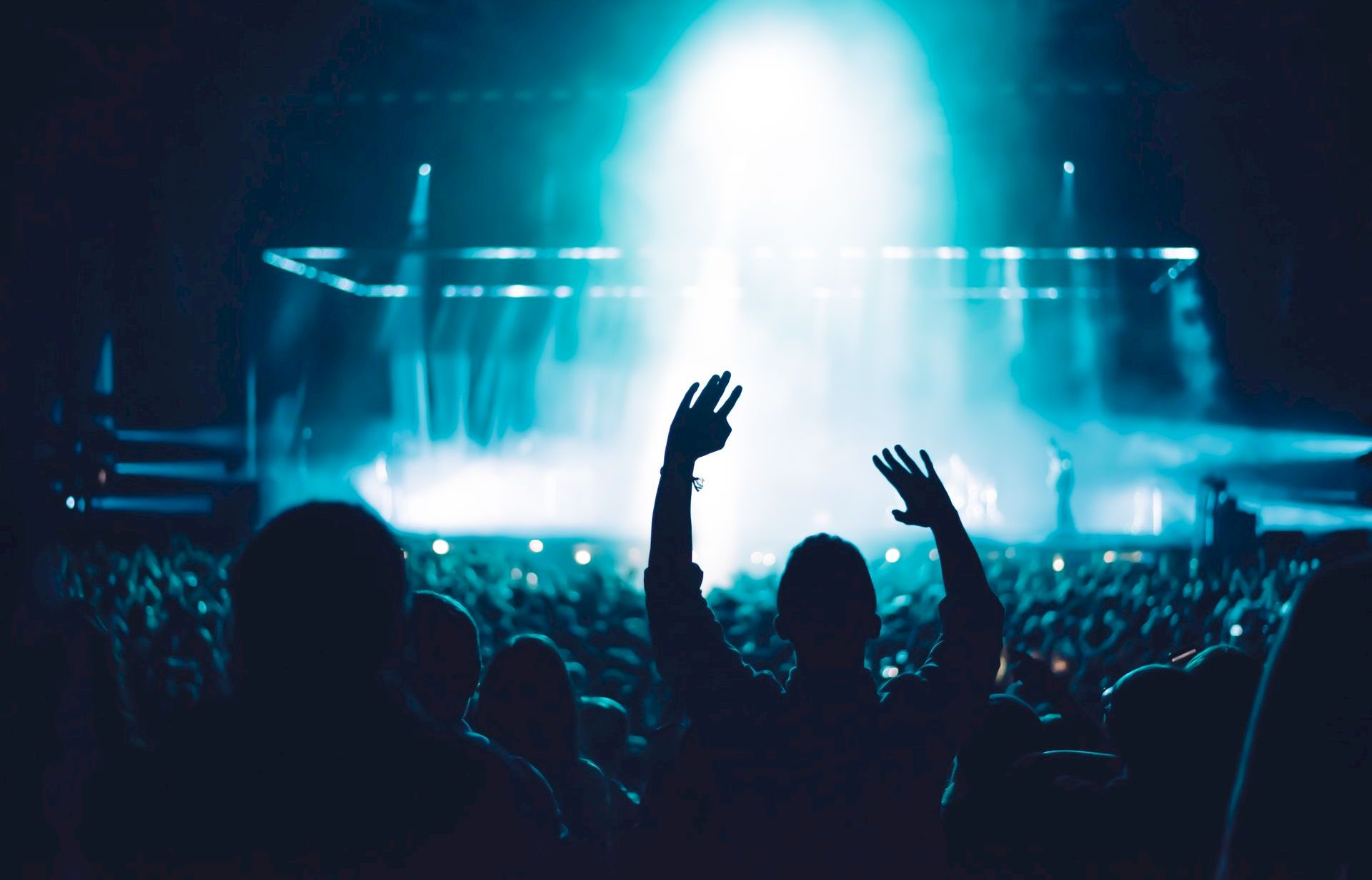 A concert crowd cheers with hands raised as a band plays under bright blue stage lights and a glowing spotlight.