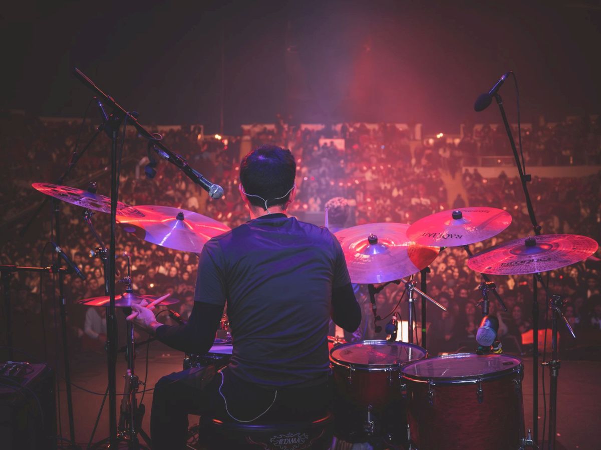 A drummer in front of colorful cymbals plays onstage while a huge crowd cheers under red and purple concert lights.