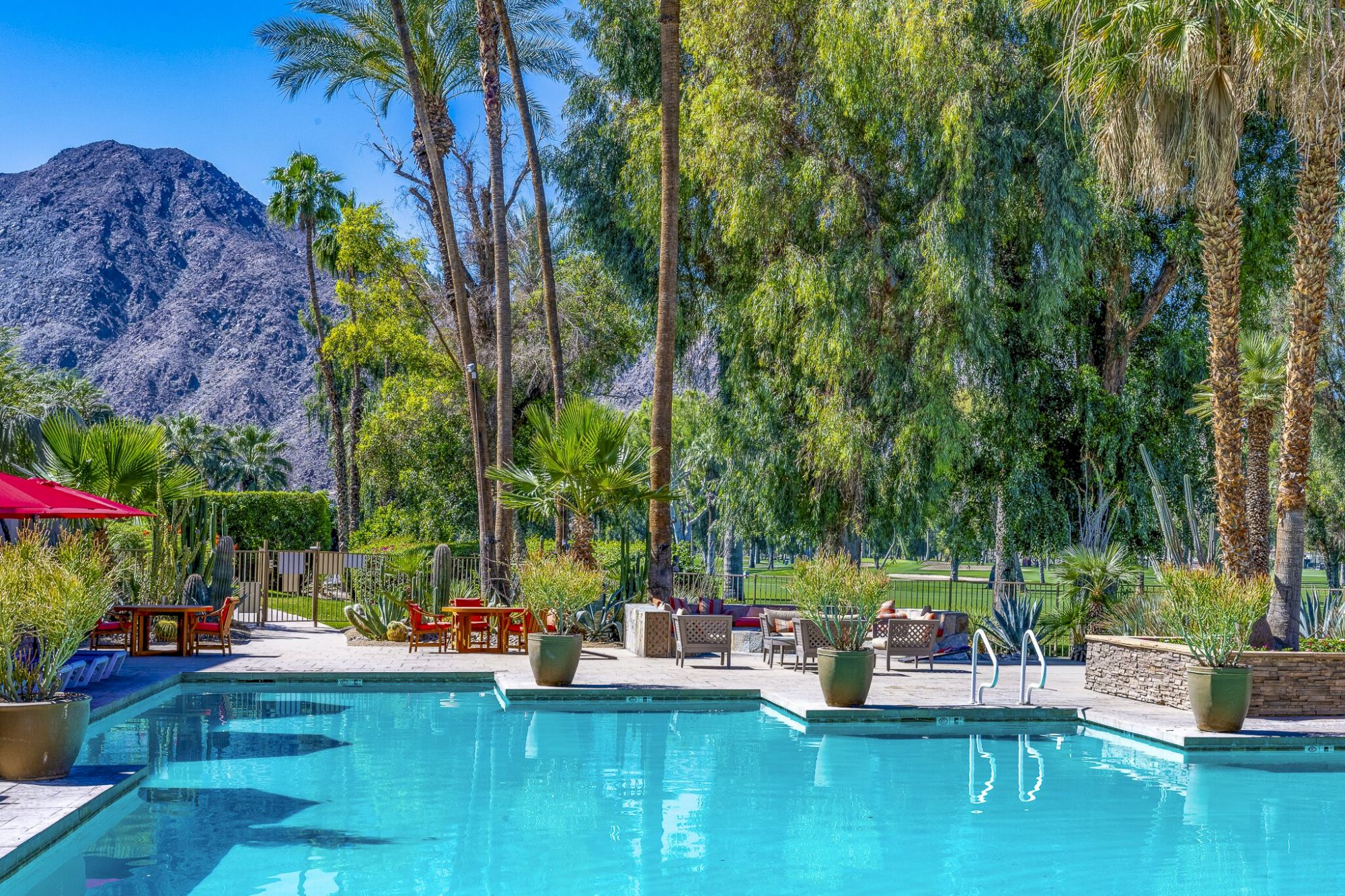 A sunny resort courtyard with a sparkling pool, lounge chairs, tall palms, cacti, and a dramatic mountain backdrop.