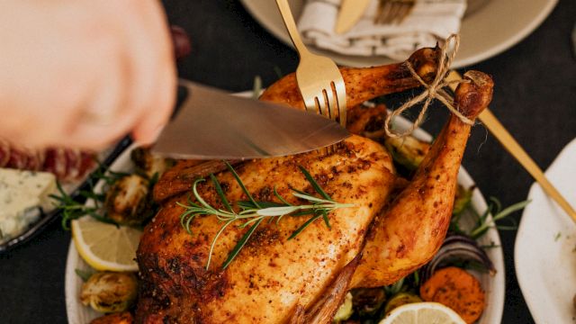 A cook is carving a roasted chicken with a fork and knife, serving it on a platter with vegetables and lemon.