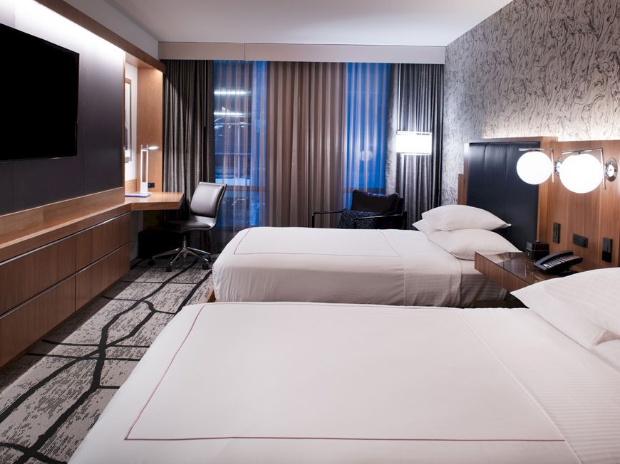 A modern hotel room with two white beds, dark wood panels, and bright wall lamps near large window curtains.