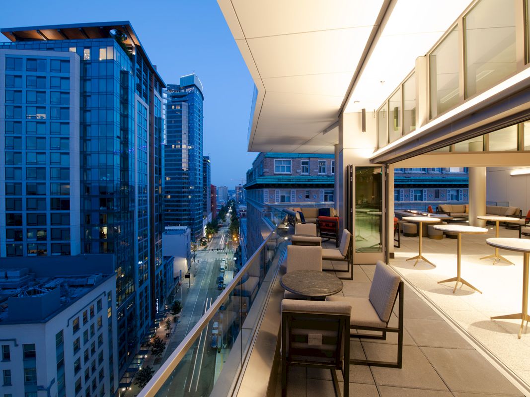 A modern high-rise rooftop terrace with lounge seating, overlooking a downtown street and tall blue glass buildings at dusk.